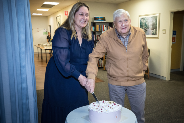 Photo of a female and male standing behind a small table with a cake on it. They are cutting the cake together.