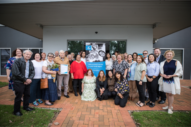 Photo of the Wesley LifeForce team gathered in front of a building. Some are holding flowers or a certificate, with a tall banner in the centre rear of the picture.