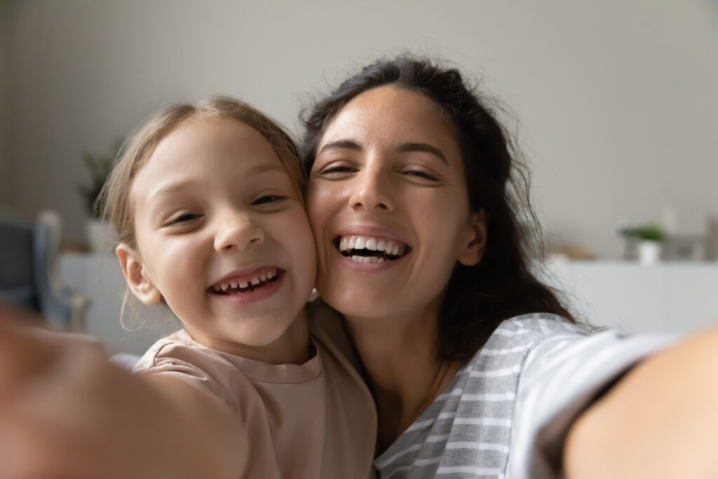 Selfie of happy young mom and sweet little daughter girl looking at webcam with toothy smile. Mother and kid taking self portrait picture on smartphone holding gadget with web camera in hand