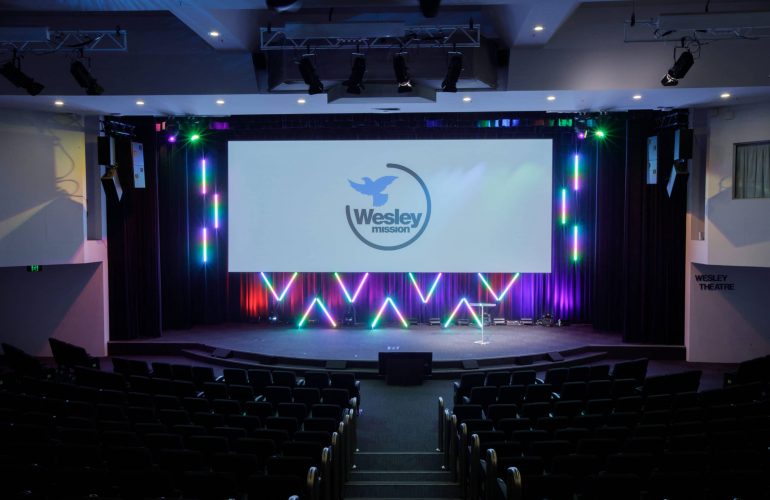 Distant front view shot of the Wesley Theatre with Wesley Mission logo showing on digital screen above the stage,