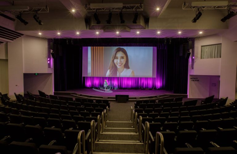 Distant front view shot of the Wesley Theatre with woman showing on digital screen above the stage